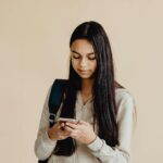 Teenage girl with long hair using a smartphone, wearing a backpack, against a beige background.